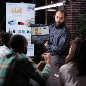 startup-entrepreneur-holding-laptop-with-sales-presentation-charts-pointing-wall-screen-tv-late-night-meeting-caucasian-man-presenting-marketing-strategy-coworkers-working-overtime