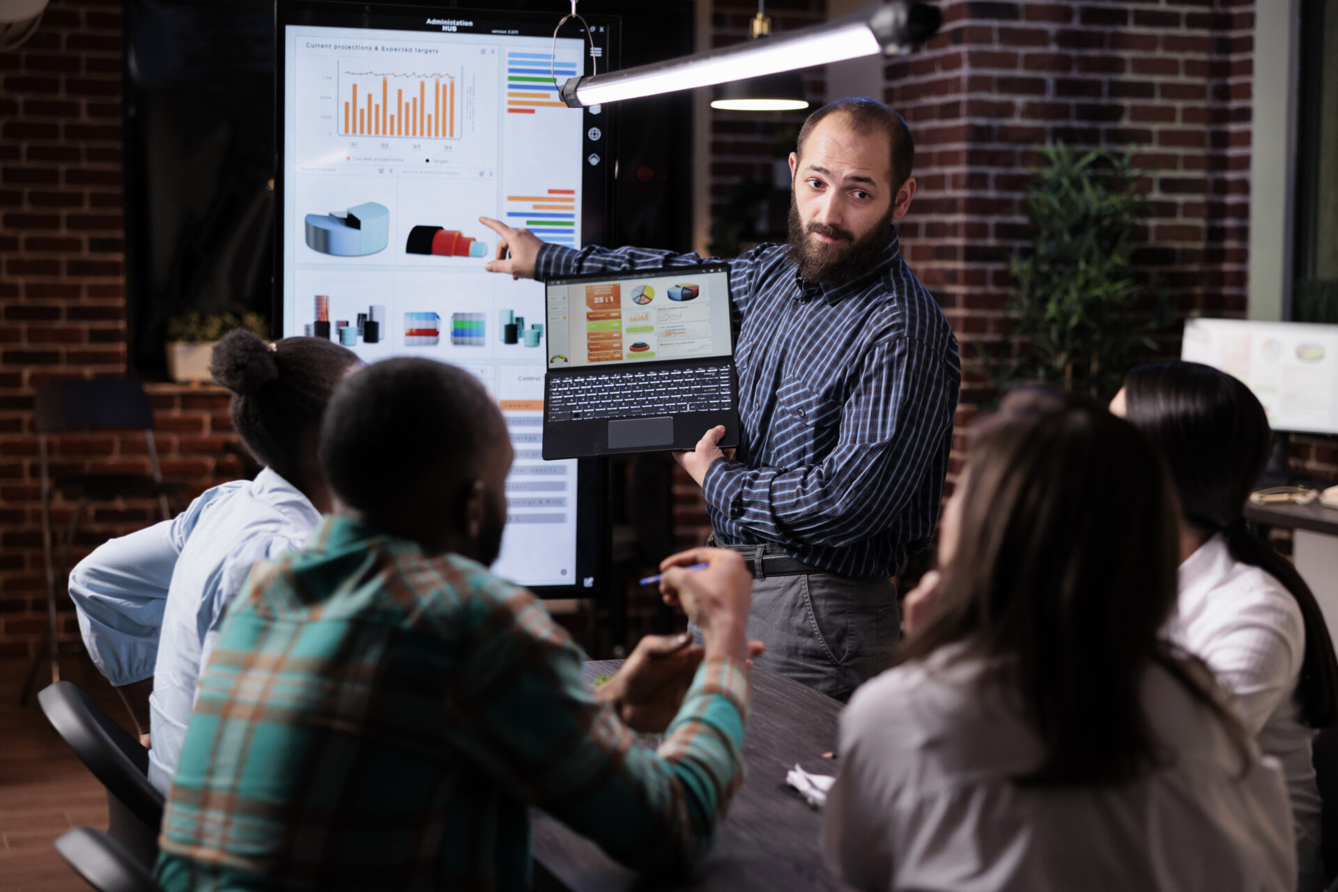 startup-entrepreneur-holding-laptop-with-sales-presentation-charts-pointing-wall-screen-tv-late-night-meeting-caucasian-man-presenting-marketing-strategy-coworkers-working-overtime